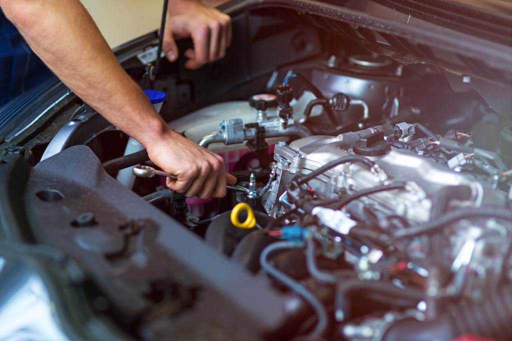 A mechanic's hand using a wrench inside a car engine bay, with various engine components visible.